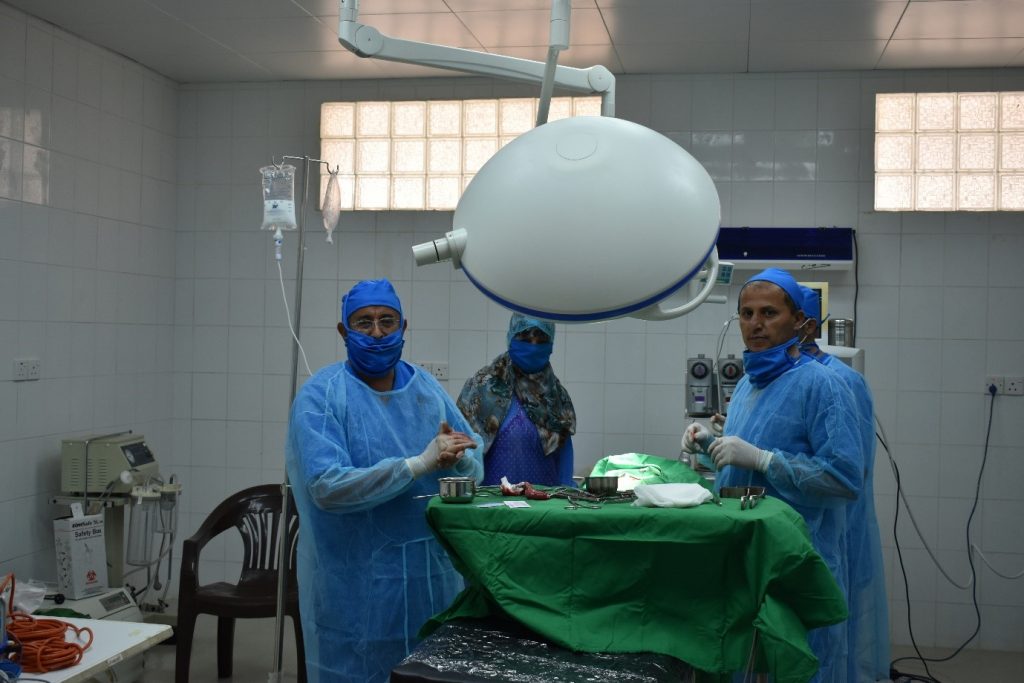Three healthcare professionals at a hospital in Yemen's Al-Soodah district, dressed in masks and surgical scrubs. gathered around an operating table, performing life-saving surgery.